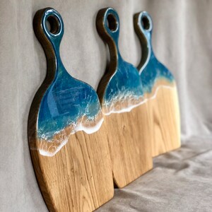 May include: Three wooden cutting boards with a blue and white resin ocean wave design. The boards have a handle with a hole for hanging.