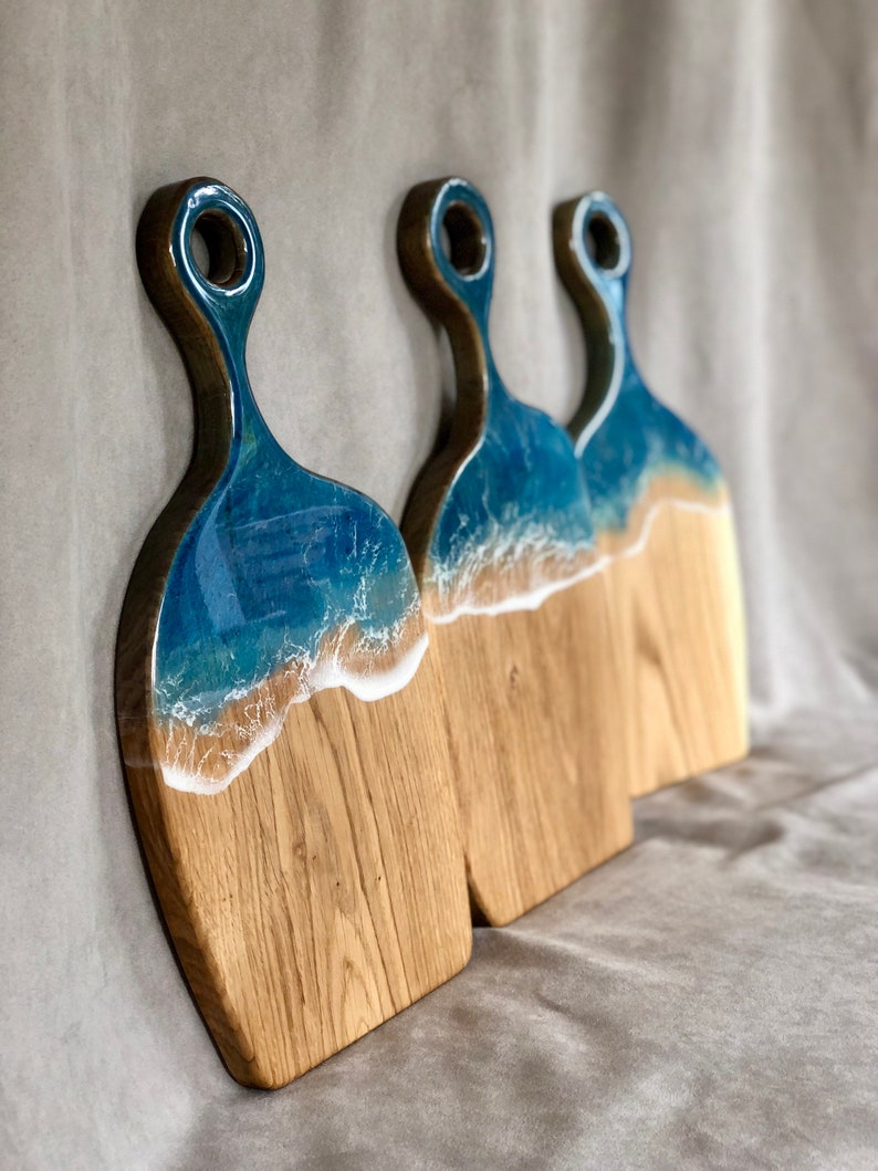 May include: Three wooden cutting boards with a blue and white resin ocean wave design. The boards have a handle with a hole for hanging.