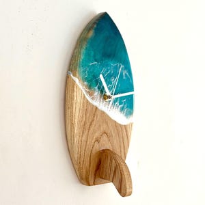 Puede incluir: Un reloj de pared con forma de tabla de surf. La parte superior es de resina turquesa con detalles blancos, imitando las olas del océano. La base es de madera. El reloj tiene manecillas blancas y una pequeña aleta.