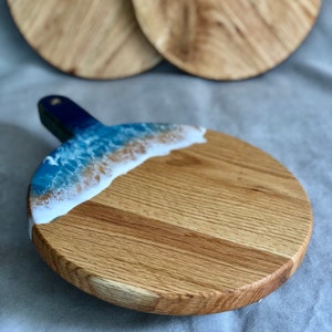 May include: A round wooden serving board with a handle, featuring a blue and white resin design resembling ocean waves. The board is made of light-coloured wood and has a natural wood grain pattern. Two additional boards are visible in the background.