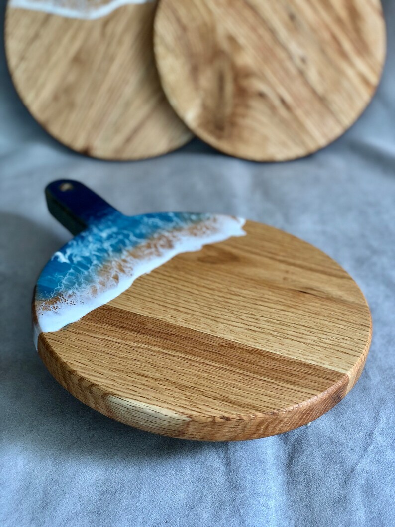 May include: A round wooden serving board with a handle, featuring a blue and white resin design resembling ocean waves. The board is made of light-coloured wood and has a natural wood grain pattern. Two additional boards are visible in the background.