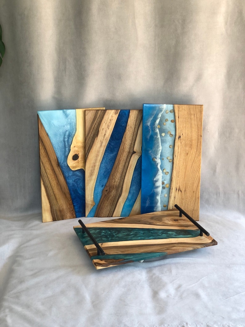 May include: Three wooden serving trays with blue and green resin inlays. The trays have a natural wood finish and black metal handles. The resin inlays have a wave-like pattern.