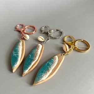 May include: Three surfboard-shaped keychains with a teal and white wave design. Each keychain has a small white seashell charm and a gold, silver, or rose gold key ring.