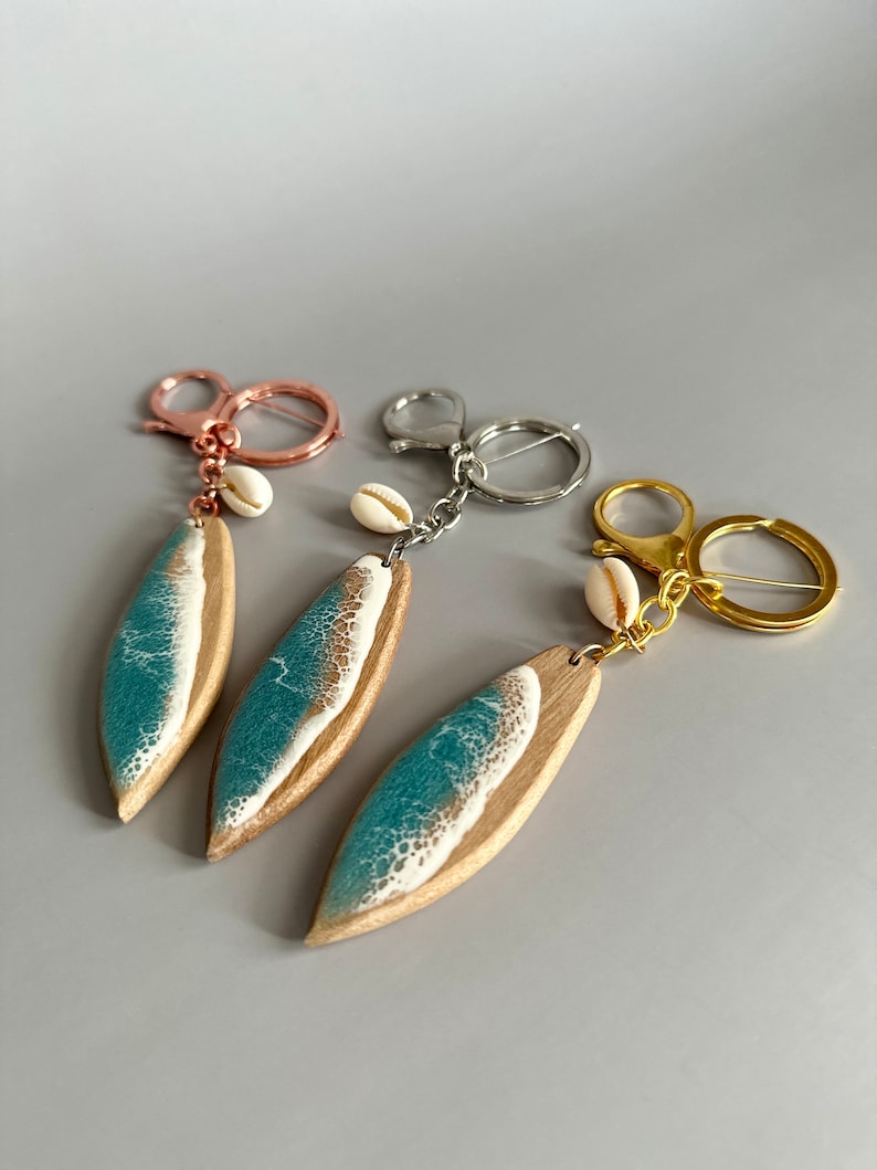 May include: Three surfboard-shaped keychains with a teal and white wave design. Each keychain has a small white seashell charm and a gold, silver, or rose gold key ring.
