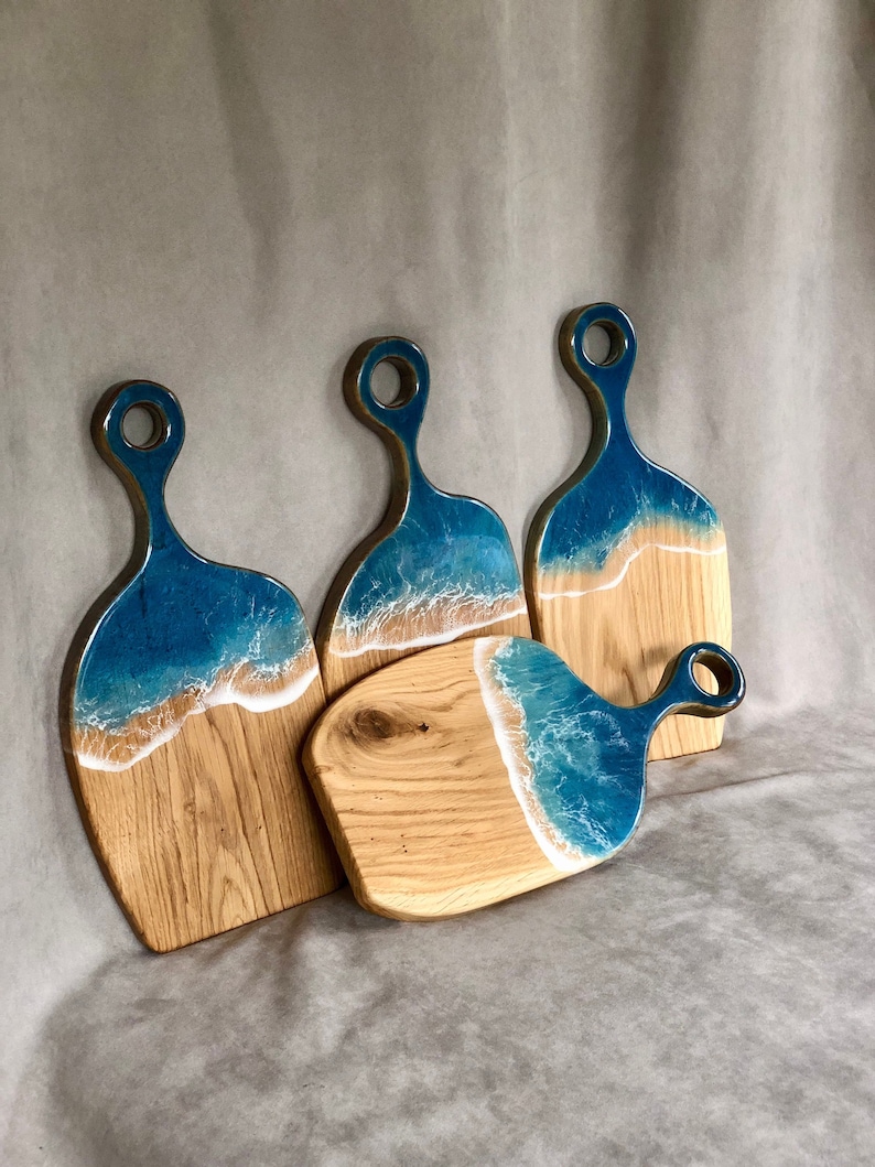 May include: Set of four wooden cutting boards with blue and white resin ocean wave designs. The boards have a rounded shape and a handle.