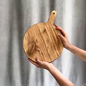 May include: A circular wooden chopping board with a handle. The board is made of light-coloured wood with a natural grain pattern. The handle has a hole for hanging. The board is held up by two hands.