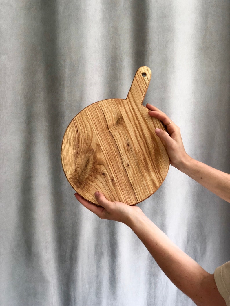 May include: A circular wooden chopping board with a handle. The board is made of light-coloured wood with a natural grain pattern. The handle has a hole for hanging. The board is held up by two hands.