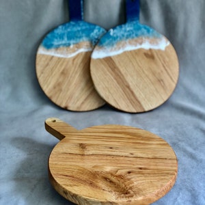 May include: Three wooden serving boards. Two boards feature a blue handle and a beach-themed design with blue resin representing the ocean and white resin for the waves. The third board is a plain wooden cutting board with a handle.