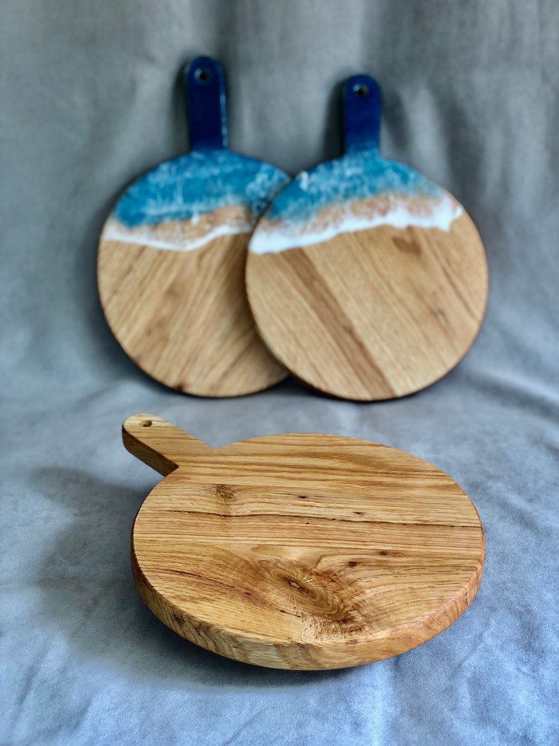 May include: Three wooden serving boards. Two boards feature a blue handle and a beach-themed design with blue resin representing the ocean and white resin for the waves. The third board is a plain wooden cutting board with a handle.