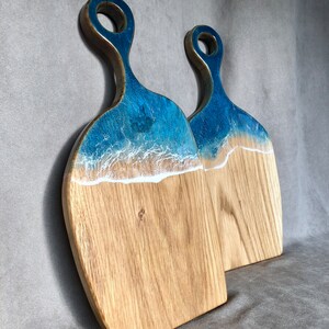 May include: Two wooden cutting boards with blue and white resin inlays that resemble ocean waves. The boards have a handle with a hole in the center.