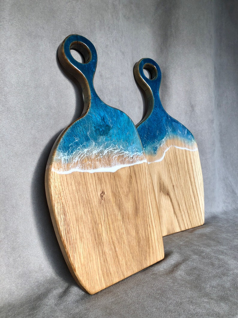 May include: Two wooden cutting boards with blue and white resin inlays that resemble ocean waves. The boards have a handle with a hole in the center.