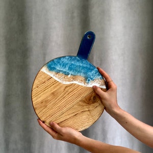 May include: Circular wooden serving board with a blue handle and a wave design. The top section has a blue and white resin wave pattern, leading to a natural wood grain base. The board is held by two hands.