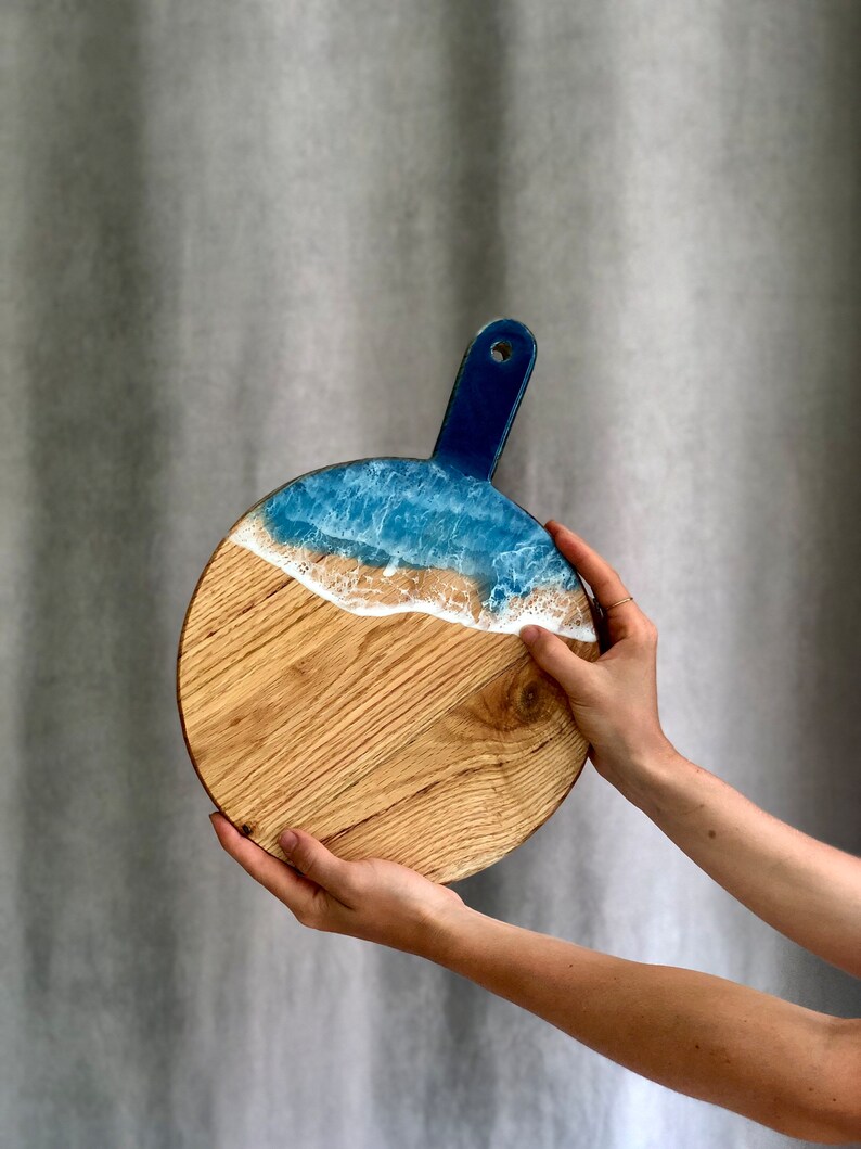 May include: Circular wooden serving board with a blue handle and a wave design. The top section has a blue and white resin wave pattern, leading to a natural wood grain base. The board is held by two hands.