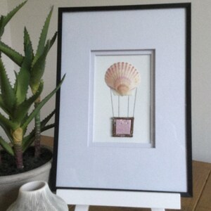 May include: A framed artwork featuring a seashell hot air balloon with a pink and white shell and a pink and silver basket. The artwork is displayed on a white easel.