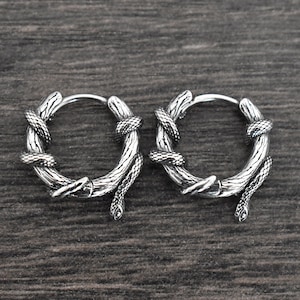 Könnte beinhalten: Ein Paar silberne Creolen mit Schlangenmuster. Die Schlangen sind ineinander verschlungen und haben eine strukturierte Oberfläche.