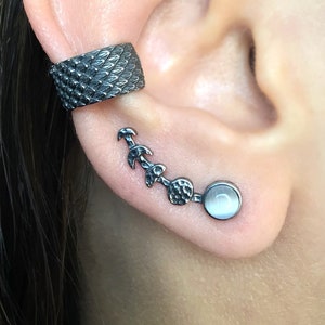 May include: A close-up of an ear adorned with two pieces of jewelry. A textured, dark gray ear cuff wraps around the upper ear. Below, a silver ear climber features a series of decorative elements and a round, white stone at the end.