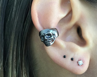 May include: A close-up of a person's ear with multiple earrings. The ear is pierced in three places. The top piercing features a silver skull-shaped earring. The middle two piercings feature small black stud earrings. The bottom piercing features a small, clear, diamond-shaped earring.