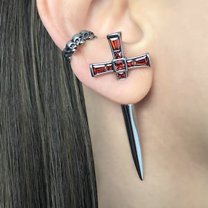 May include: A pair of earrings featuring a silver cross with red gemstones and a silver dagger-shaped earring.