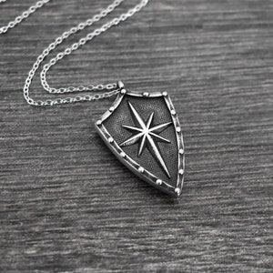 以下が含まれることがあります： 星形のデザインが施された盾のペンダントが付いたシルバーネックレス。盾はテクスチャ加工が施され、ビーズの縁取りがされています。