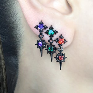 May include: Three pairs of black dangle earrings with starburst designs and colorful crystal accents. The earrings feature a small crystal at the top and a larger crystal at the bottom of the dangle.