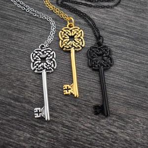 May include: Three key pendant necklaces in silver, gold, and black. Each key has a decorative head and hangs from a matching chain. The keys are displayed on a dark gray wooden surface.