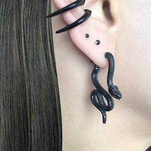 Peut inclure: Boucles d'oreilles en forme de serpent noir avec une queue enroulée. Les boucles d'oreilles sont en matériau noir lisse et sont portées au lobe de l'oreille.