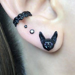 May include: Black cat head stud earrings with purple gem eyes. A black skull ear cuff and two small black gem studs are also visible. The jewelry is worn on an ear.