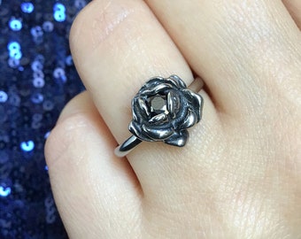 Könnte beinhalten: Ein silberner Ring mit einem Rosenmotiv. Die Rose ist aus Metall und hat eine dunkelgraue Oberfläche. Der Ring wird an einem Finger getragen.