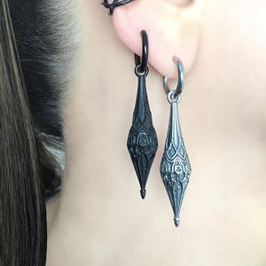 May include: A pair of black dangle earrings with intricate designs, one with a hoop and the other with a curved bar, worn with a black ear cuff.