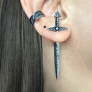 May include: A silver sword earring with a black gem in the center. The earring is worn in the upper lobe of an ear. A silver ring with a Celtic knot design is worn in the cartilage of the same ear.