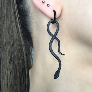 Könnte beinhalten: Schwarzer Schlangen-Ohrhänger mit einem Ring oben. Der Ohrring ist aus Metall und hat eine strukturierte Oberfläche. Die Schlange ist zusammengerollt und hat einen kleinen Kopf. Der Ohrring hängt am Ohr einer Person.
