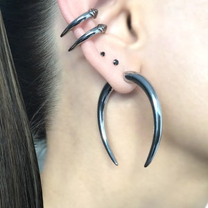 May include: A person with long brown hair wearing multiple silver ear piercings. The piercings include two small studs and a large curved hoop.