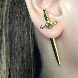 May include: Gold sword earrings with a green gemstone. The sword is a classic design with a pointed tip and a cross guard.