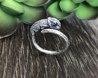 May include: A silver-toned ring in the shape of a chameleon, with a textured body and a blue gemstone eye. The ring wraps around the finger, creating a unique and artistic design. The ring is set against a dark wood surface.