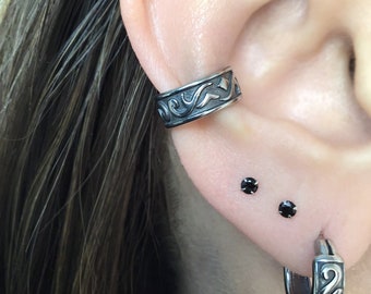 May include: A close-up of a person's ear with three earrings. The top earring is a silver ear cuff with a black floral design. The bottom two earrings are small black studs.