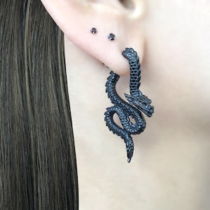 Peut inclure: Boucles d'oreilles en forme de dragon noir. Les boucles d'oreilles sont en métal et présentent un design détaillé. Les boucles d'oreilles pendent du lobe de l'oreille.