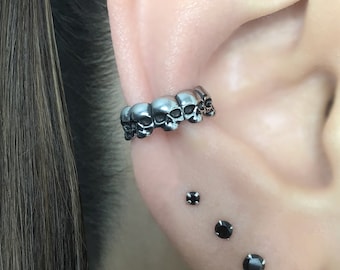 May include: A silver skull ear cuff with five skulls in a row. Three black gemstone studs are in the earlobe.