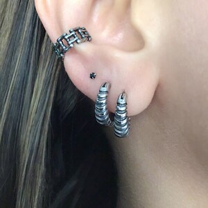 May include: A person's ear with three silver earrings. The top earring is a chain link design, the middle earring is a small black stud, and the bottom two earrings are silver hoops with a textured design.