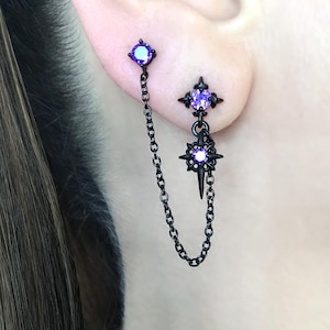 May include: Black double-piercing earrings with purple gemstones. One earring features a square gem and a chain connecting to a starburst design with a central gem and a dangling spike. The other earring has a square gem.