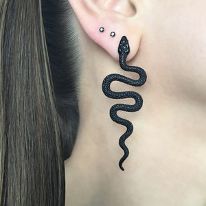 Könnte beinhalten: Ein schwarzer Schlangenohrring mit einer strukturierten Oberfläche. Die Schlange ist aufgerollt und hat einen spitzen Kopf. Der Ohrring wird am Ohrläppchen getragen.