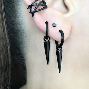 May include: An ear featuring black jewellery. A black ear cuff encircles the upper ear, complemented by two hoop earrings with cone pendants. A small black stud earring is also present. The jewellery has a gothic aesthetic.