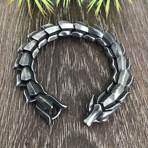 Puede incluir: Una pulsera plateada con un dise&ntilde;o de drag&oacute;n. La pulsera est&aacute; hecha de metal y tiene una superficie texturizada.