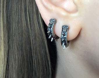 May include: A pair of silver hoop earrings with a skull design and spikes. The earrings are worn in the earlobes.