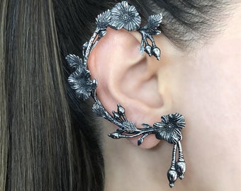 Puede incluir: Un pendiente de oreja de plata con un diseño floral. El pendiente tiene forma de rama con hojas y flores. Las flores son detalladas y tienen un aspecto realista.