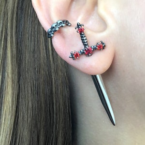 May include: A pair of black ear cuffs with red gemstone accents. One cuff is a skull design, the other is a sword design with a long, pointed dagger hanging from it.