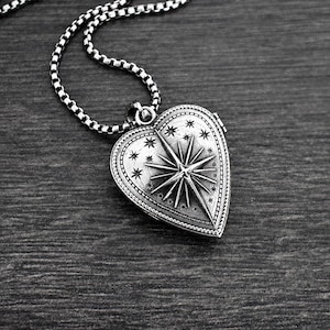 May include: A silver-toned heart-shaped locket pendant suspended from a matching box chain. The locket features an embossed North Star design surrounded by small engraved stars, with a beaded border detail along the outer edge of the heart.
