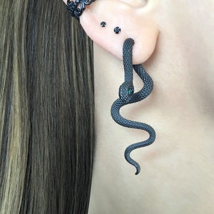 May include: Black snake ear cuff with a green gemstone eye. The snake is curled around the ear and has a textured surface.