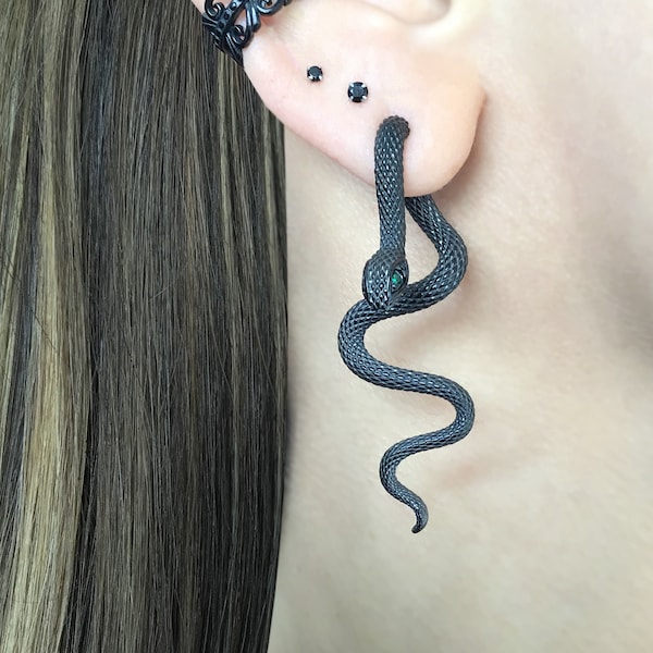 Può includere: Orecchino a forma di serpente nero con un occhio di pietra preziosa verde. Il serpente è avvolto attorno all'orecchio e ha una superficie testurizzata.