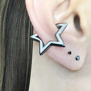 Könnte beinhalten: Ein silberfarbener sternförmiger Ohrschmuck wird am Ohr getragen. Zwei kleine schwarze Ohrstecker sind ebenfalls sichtbar. Der Ohrschmuck hat ein Zickzack-Design und besteht aus glänzendem Metall.