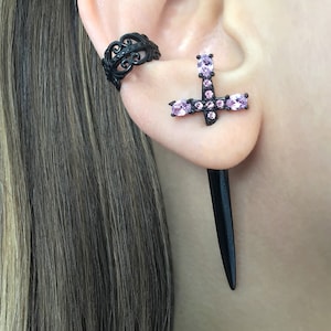 May include: Black ear cuff with a delicate filigree design and a black cross earring with pink rhinestones. The cross earring is attached to a long, black dagger-shaped dangle.